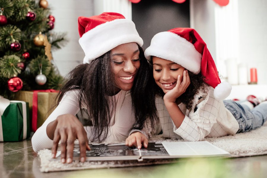 Woman and girl lying on stomachs on floor, wearing Santa hats, looking at photo album together near Christmas tree