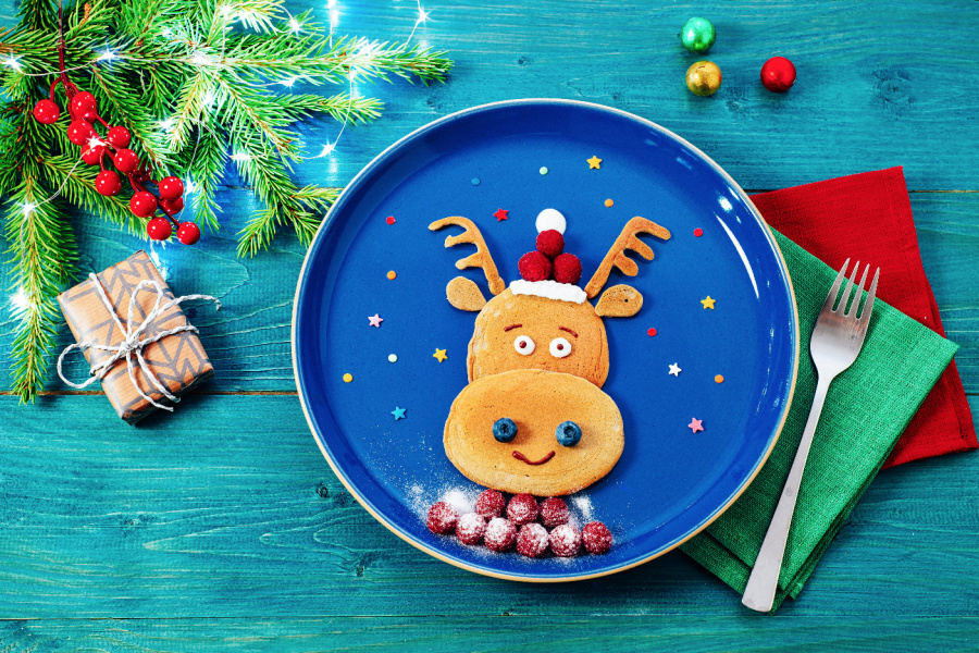 Pancake in the shape of a reindeer face with fruit on a blue plate with Christmas decor on the table. 